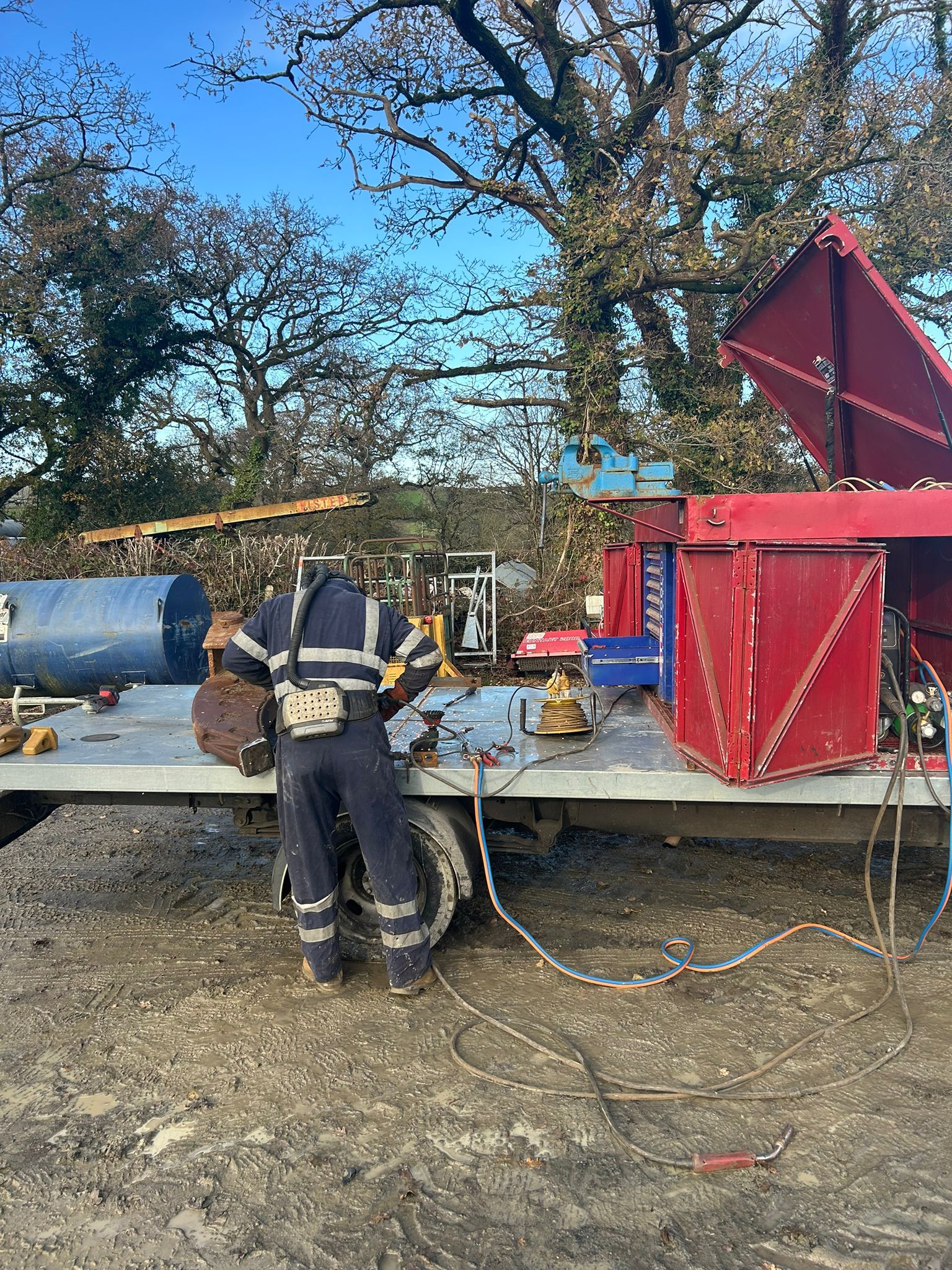A person in work attire stands by a flatbed truck with open red toolboxes and equipment, ready for mobile welding tasks. Trees and various construction materials frame the work site on a clear day.