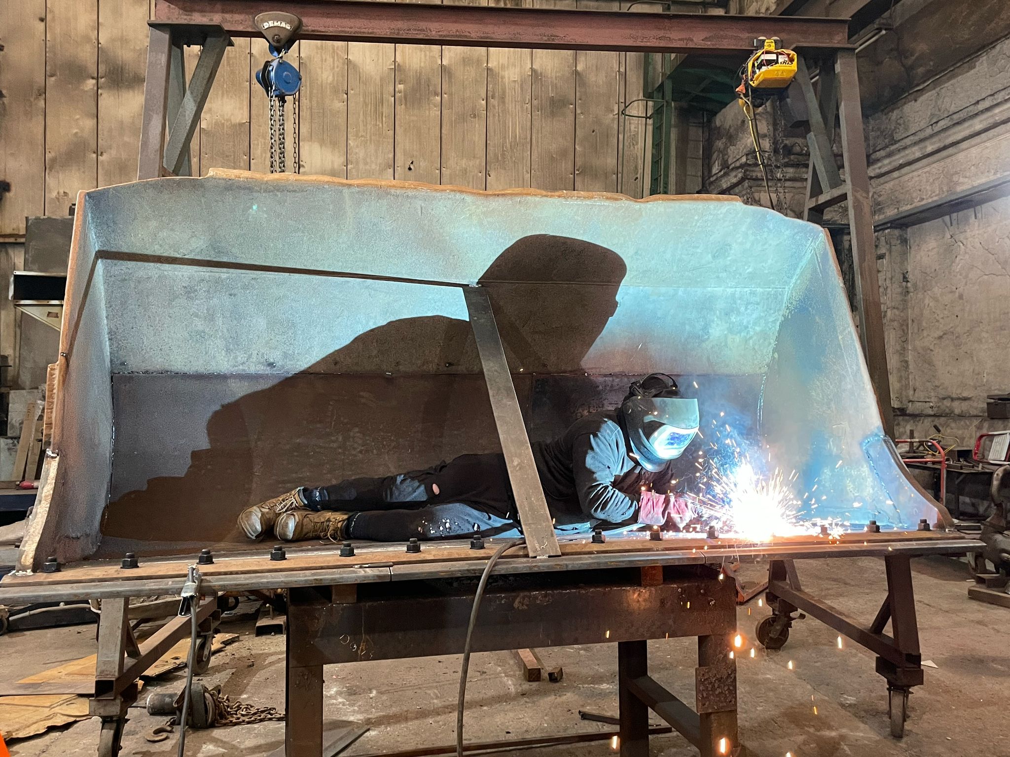 A welder, specializing in plant repair, lies inside a large metal structure, welding torch in hand and bright sparks flying. The industrial workshop has wooden walls, metal beams, and a hoist in the backdrop. Clad in safety gear including a helmet, the worker is focused on precision and safety.