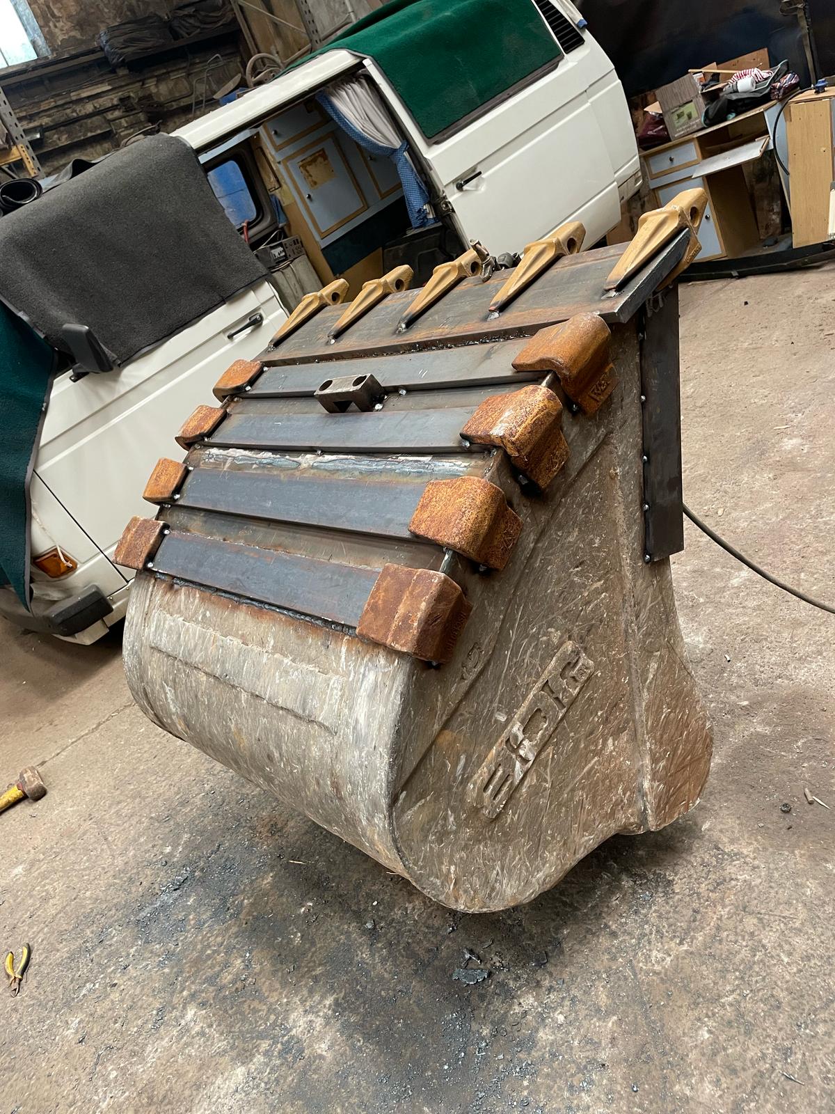 A large, weathered bucket with metal edges and several wooden handles stands on a concrete floor, hinting at the recent visit of a mobile welder. A white van and various garage items linger in the background.