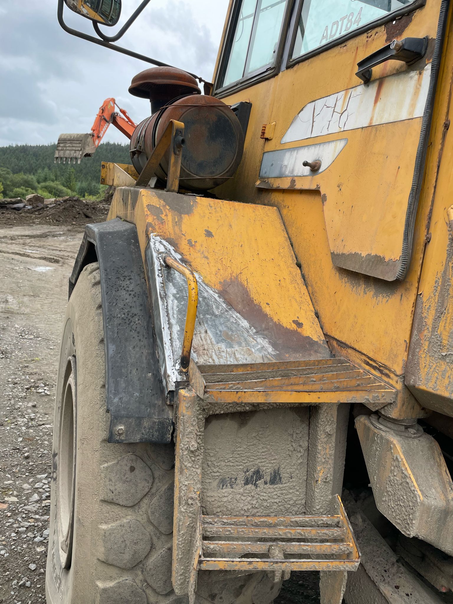 A close-up of a large, yellow, mud-splattered construction vehicle with visible wheel, step, and rear section showcases the rugged reliability essential for plant repair. In the background, there's equipment and a forested area under a cloudy sky.