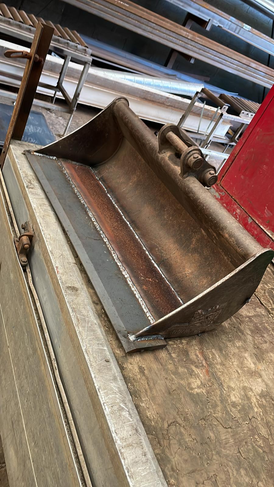 A large metal excavator bucket with a rusty interior rests on a wooden surface in an industrial setting, ready for MOT welding. Nearby, metal beams and pipes are stacked, awaiting plant repair by the skilled mobile welder.