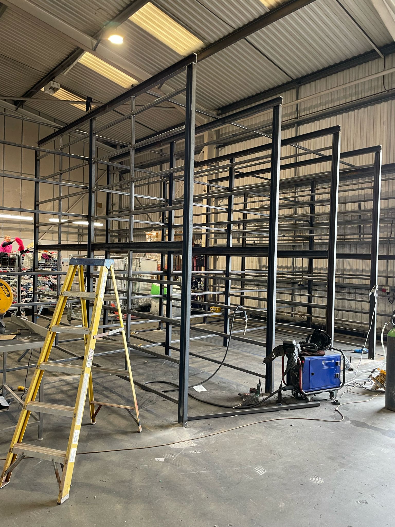 In the industrial warehouse, a mobile welder is at work as a large metal shelving unit is being assembled. A yellow ladder stands ready, surrounded by various tools and equipment on the floor, while overhead lights cast a bright glow over the diligent scene.