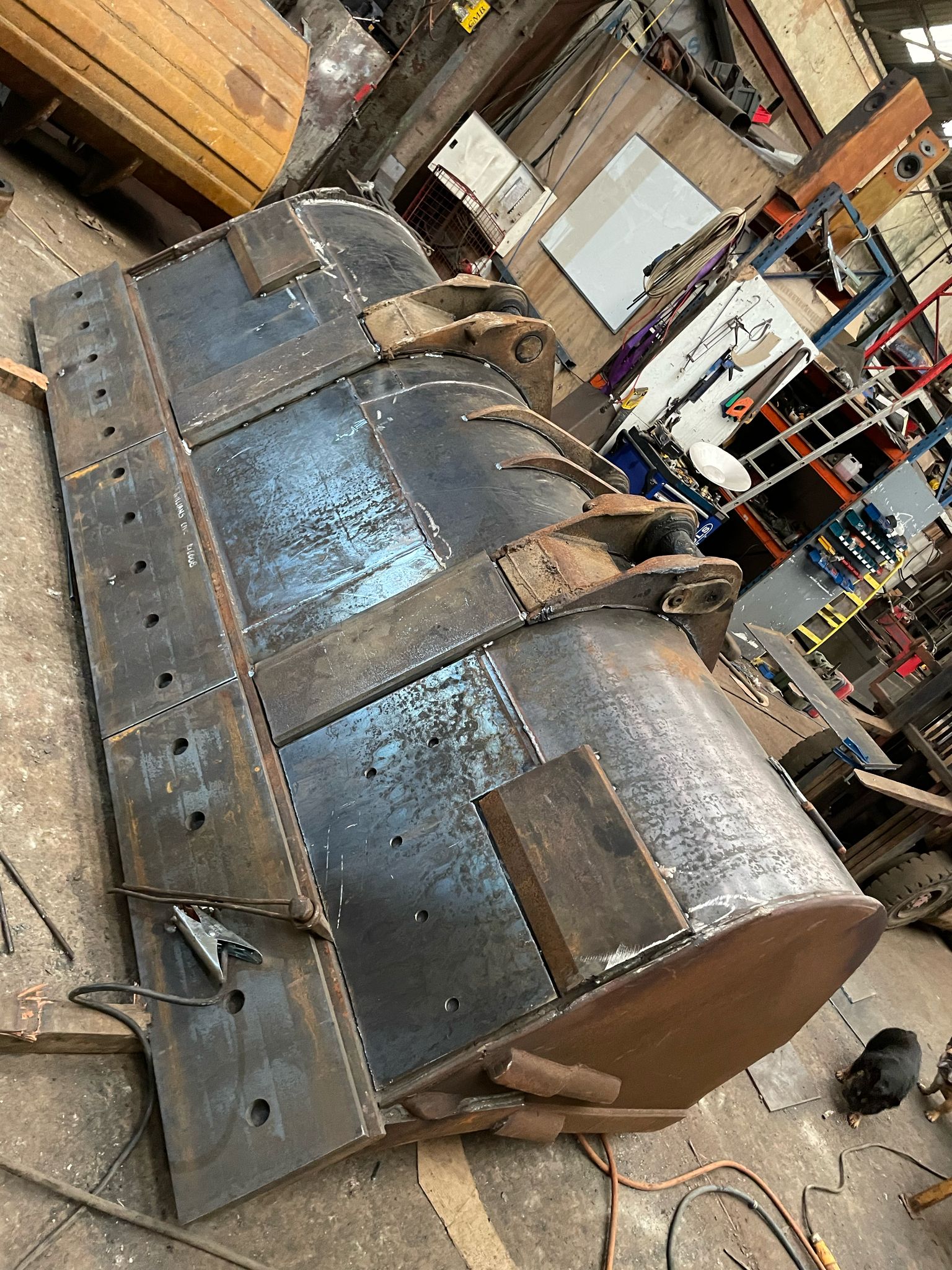 A large metal industrial attachment, potentially part of heavy machinery, rests on a workshop floor. Scattered tools hint at recent plant repair work, while the backdrop reveals a partially visible wooden table surrounded by an array of equipment.