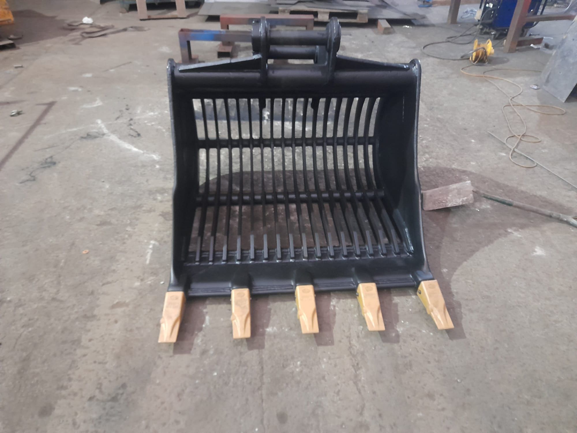 A metal excavator bucket with five teeth and slatted sides sits on a concrete floor in a workshop. Scattered in the background are tools and equipment, essential for plant repair and mobile welding tasks, hinting at the industrious activities undertaken here.