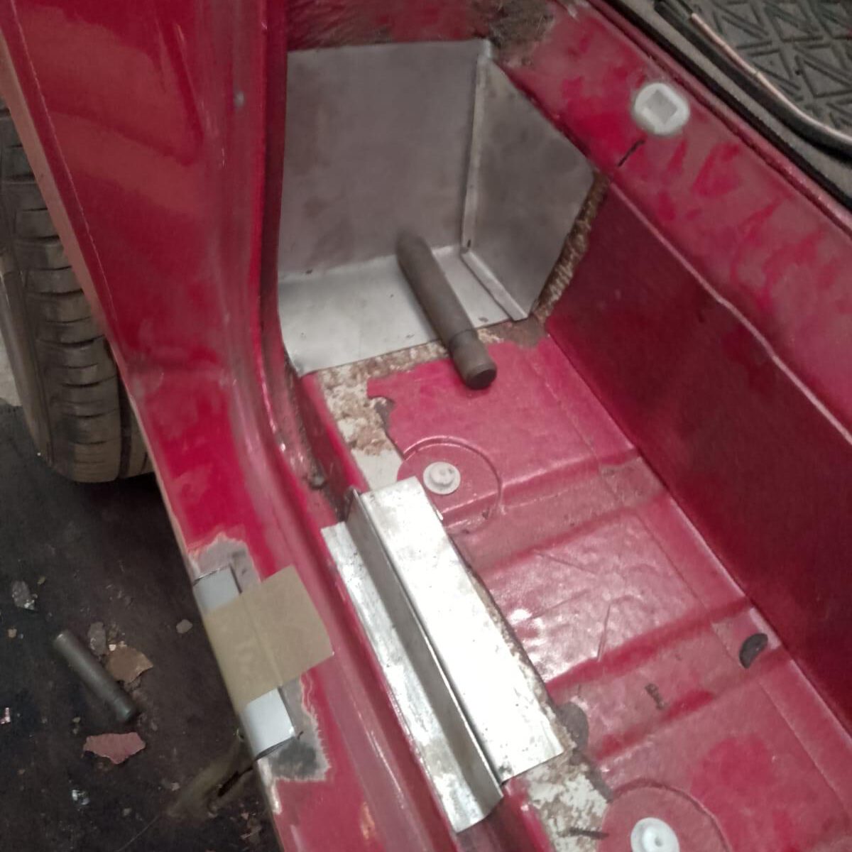 Interior view of a car's red-painted floor, showing visible wear with rust spots and metal reinforcements, ideal for an expert in MOT welding. A small pipe protrudes near the corner. One wheel and part of the door frame are seen on the left side.