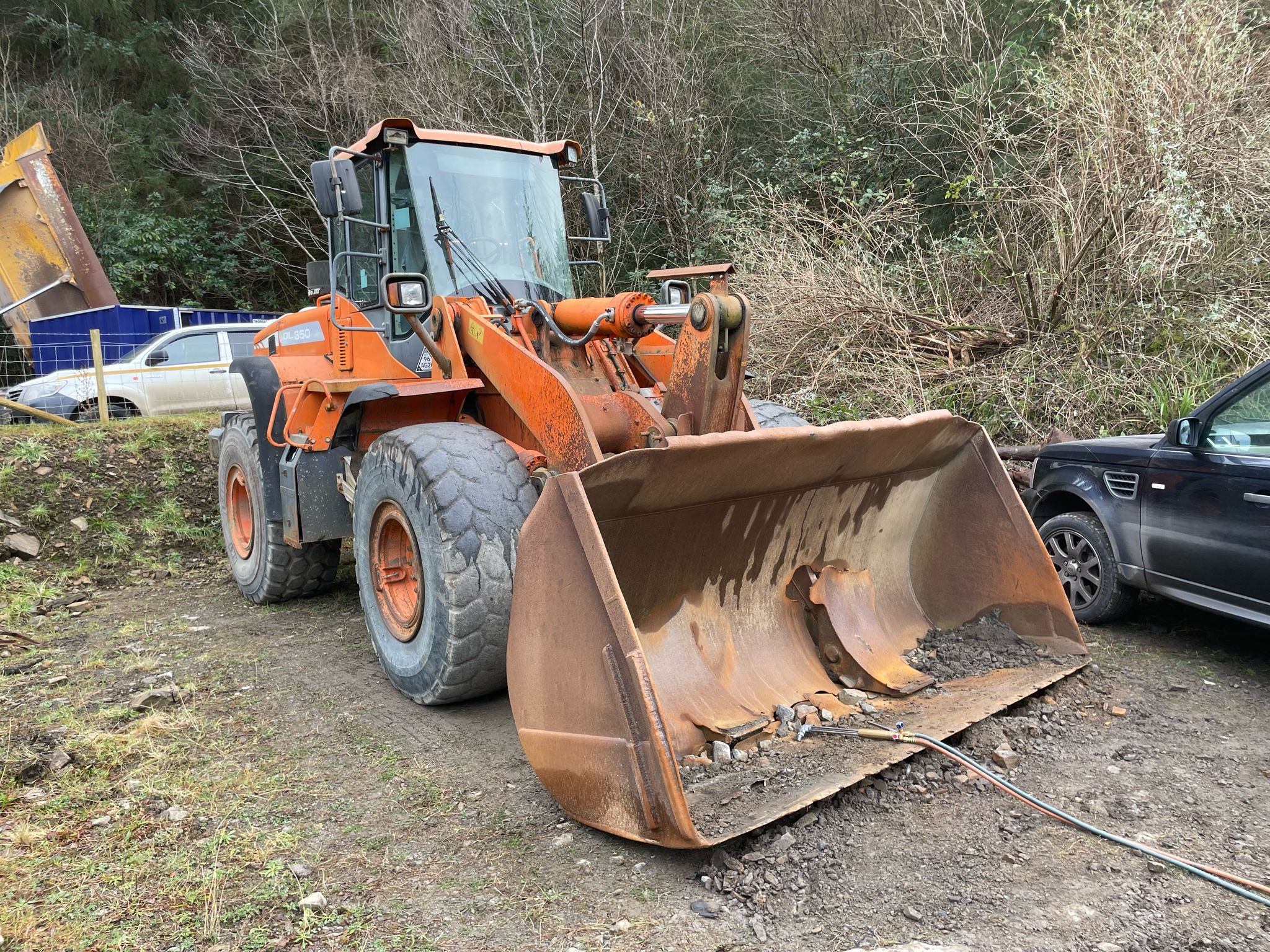 An orange front-end loader with a rusted bucket sits in solitude on a dirt path near a forest. Next to it, a black vehicle stands watch, while the fence and trees behind speak of quiet maintenance days, hinting at the touch of MOT Welding or even Plant Repair expertise.