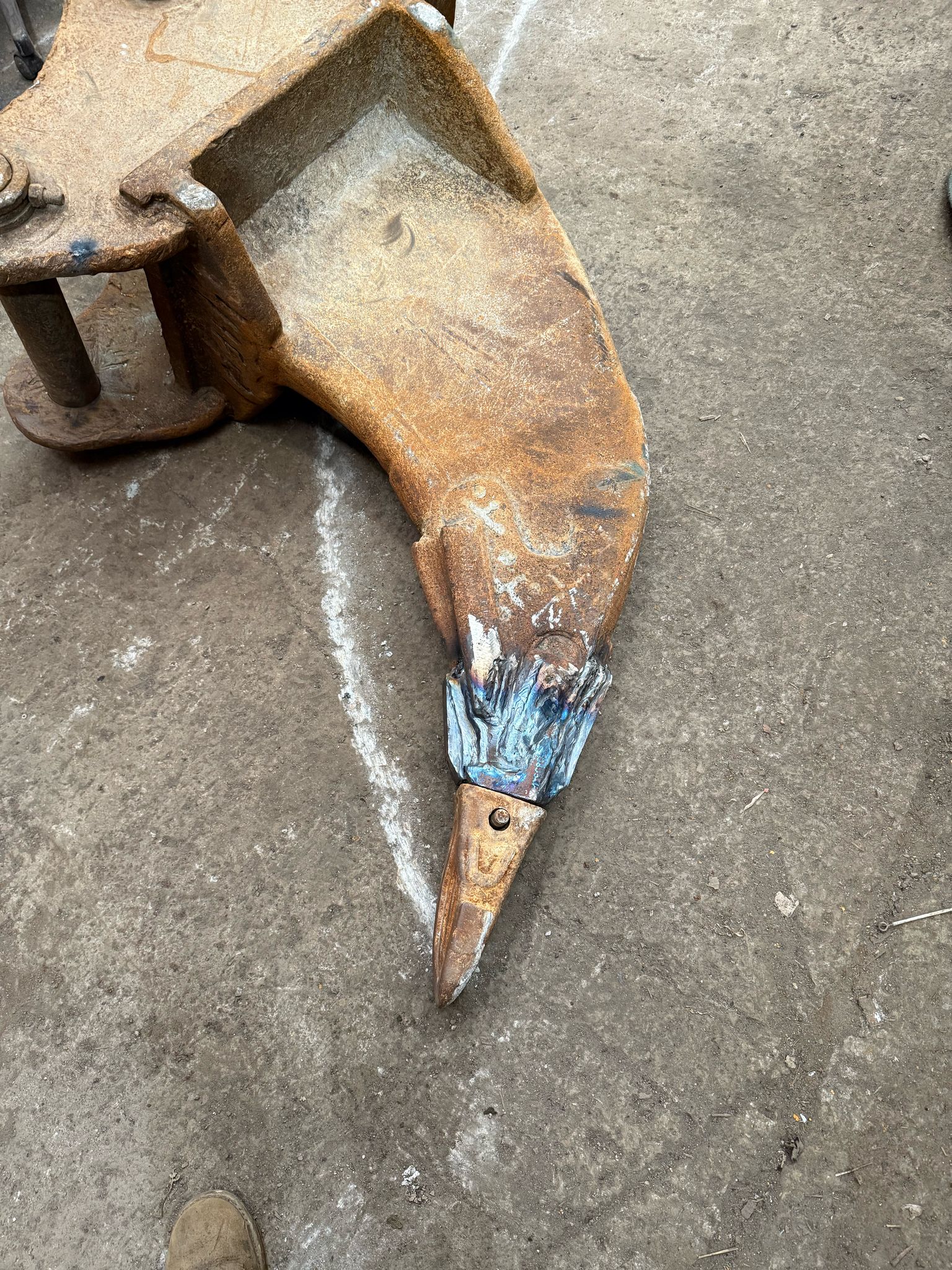 A close-up of a large, rusted metal tool on the ground, resembling an excavator tooth, evokes images of plant repair. The pointed end is worn and has blue paint remnants. The concrete surface beneath shows a faint white line, hinting at where mobile welders once restored its strength.