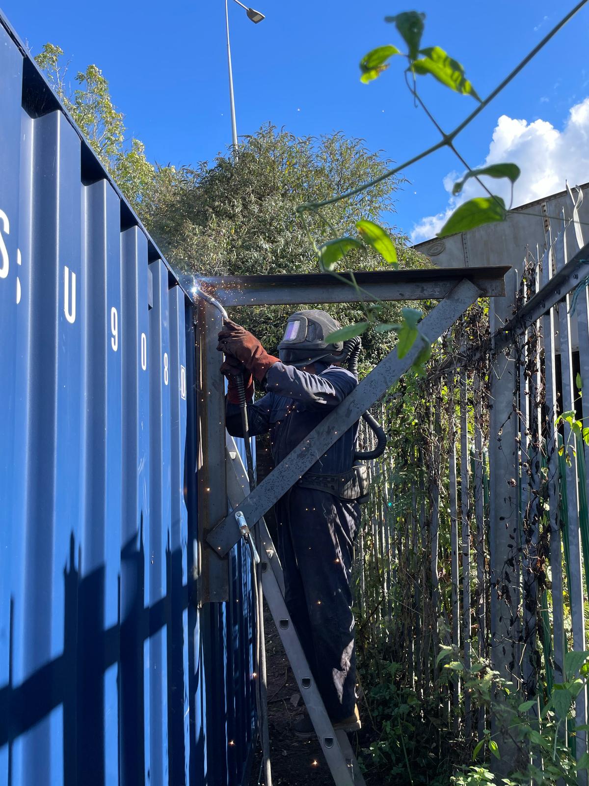 A mobile welder in protective gear and helmet is expertly welding a metal structure on the side of a blue shipping container. Nearby, green plants flourish beside a metal fence, all under a bright blue sky dotted with white clouds.