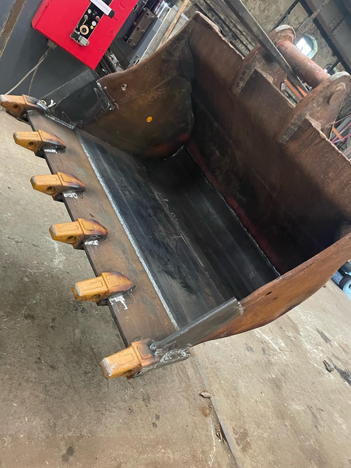 Close-up of an excavator bucket with sturdy teeth visible, poised on a workshop floor where plant repair takes place. The bucket is angled upward, and a red tool chest, used by the mobile welder for MOT welding tasks, is partially visible in the background.