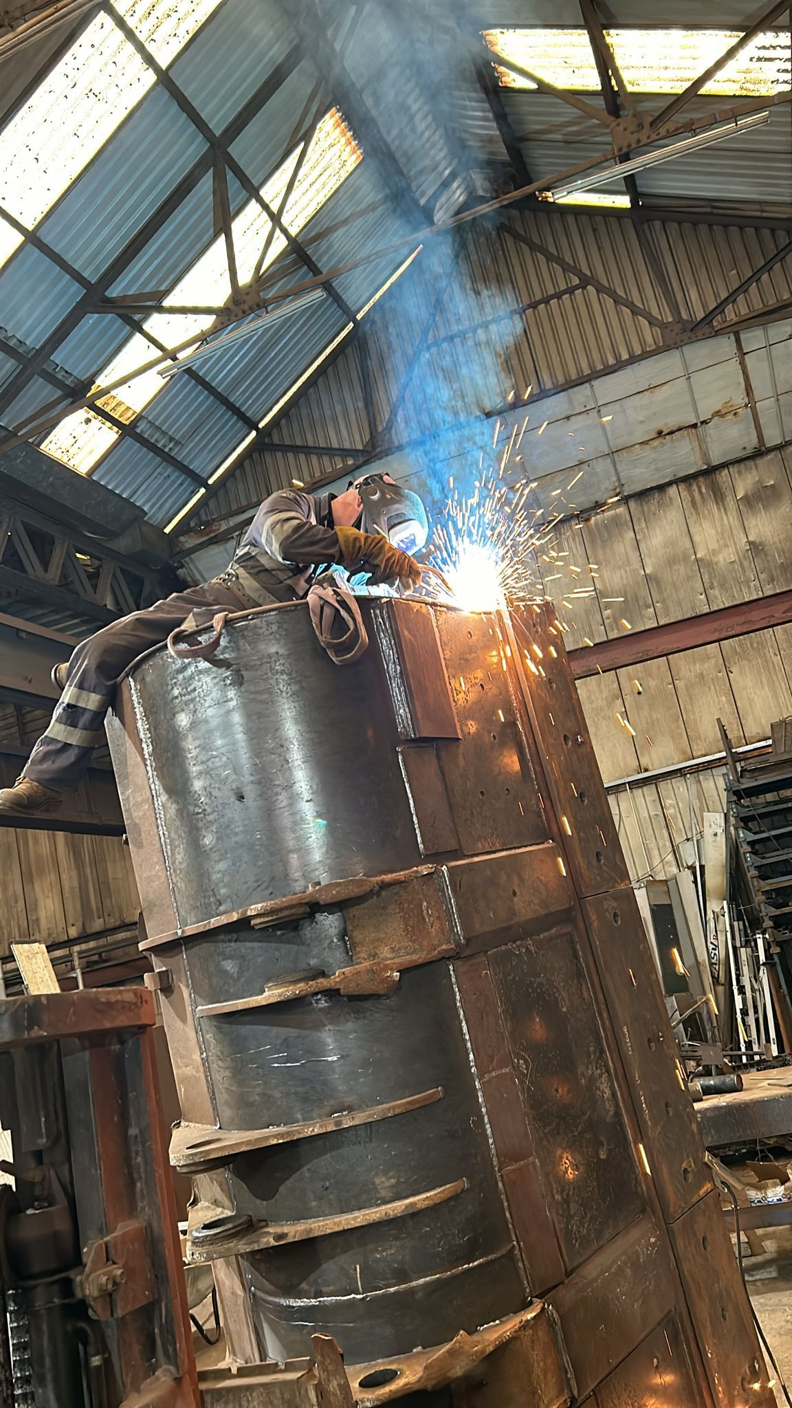 A mobile welder is busy welding on top of a large metal structure inside an industrial warehouse. Sparks fly, illuminating the scene filled with metal beams and a corrugated roof. The worker, focused on plant repair, diligently wears safety gear.