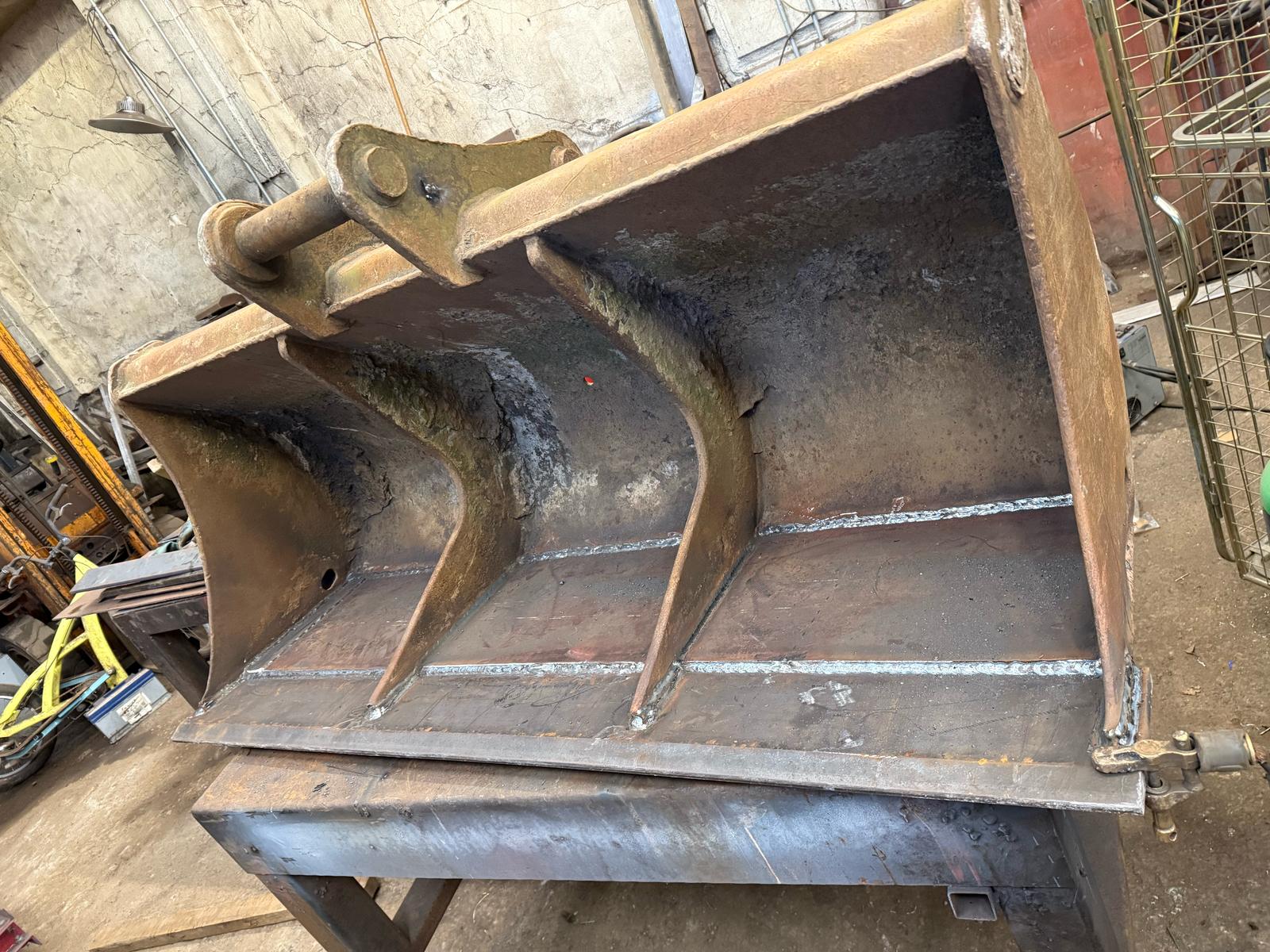 A large, rusted metal excavator bucket sits on a platform in a bustling plant repair workshop. The bucket features three prominent teeth and welded seams, showcasing expert MOT welding work. The background includes various tools and equipment, with a metal fence on the right.