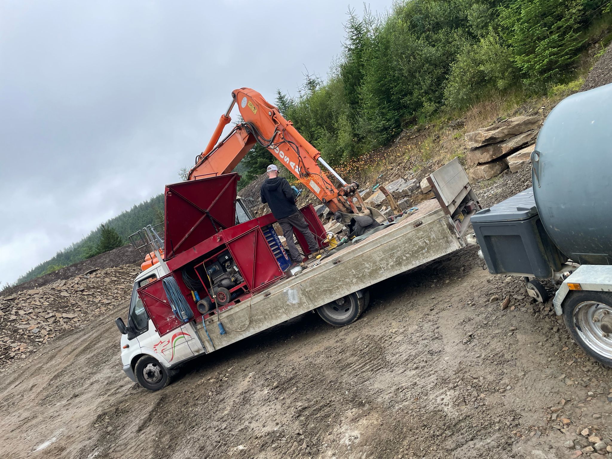 A person stands on a flatbed truck, operating a red machinery unit next to an orange excavator. The scene, which involves plant repair and mobile welding tasks, unfolds on a muddy construction site surrounded by trees under a cloudy sky.