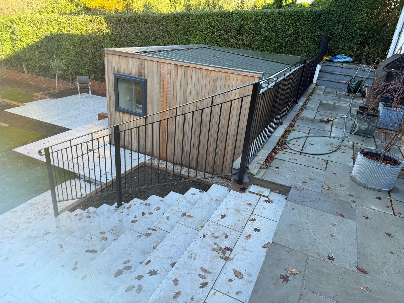 View of a backyard with a wooden shed next to a patio, where a mobile welder has left their mark. Stone steps with a black metal railing lead down to the patio. A garden hose is coiled on the right, and leaves are scattered on the steps.