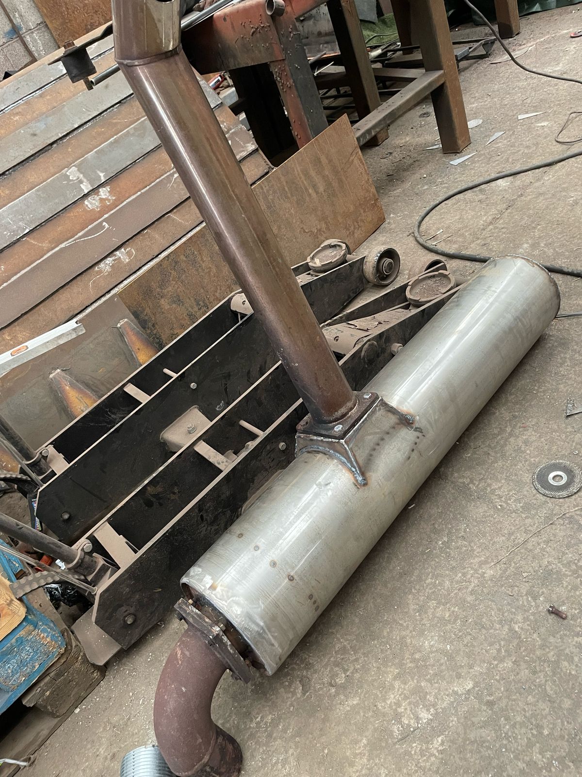 A metal exhaust pipe connected to a large cylindrical muffler rests on the workshop floor. Amidst the array of tools and equipment, hinting at a space for MOT welding and metal fabrication, some plywood leans against the wall, completing this industrious scene.