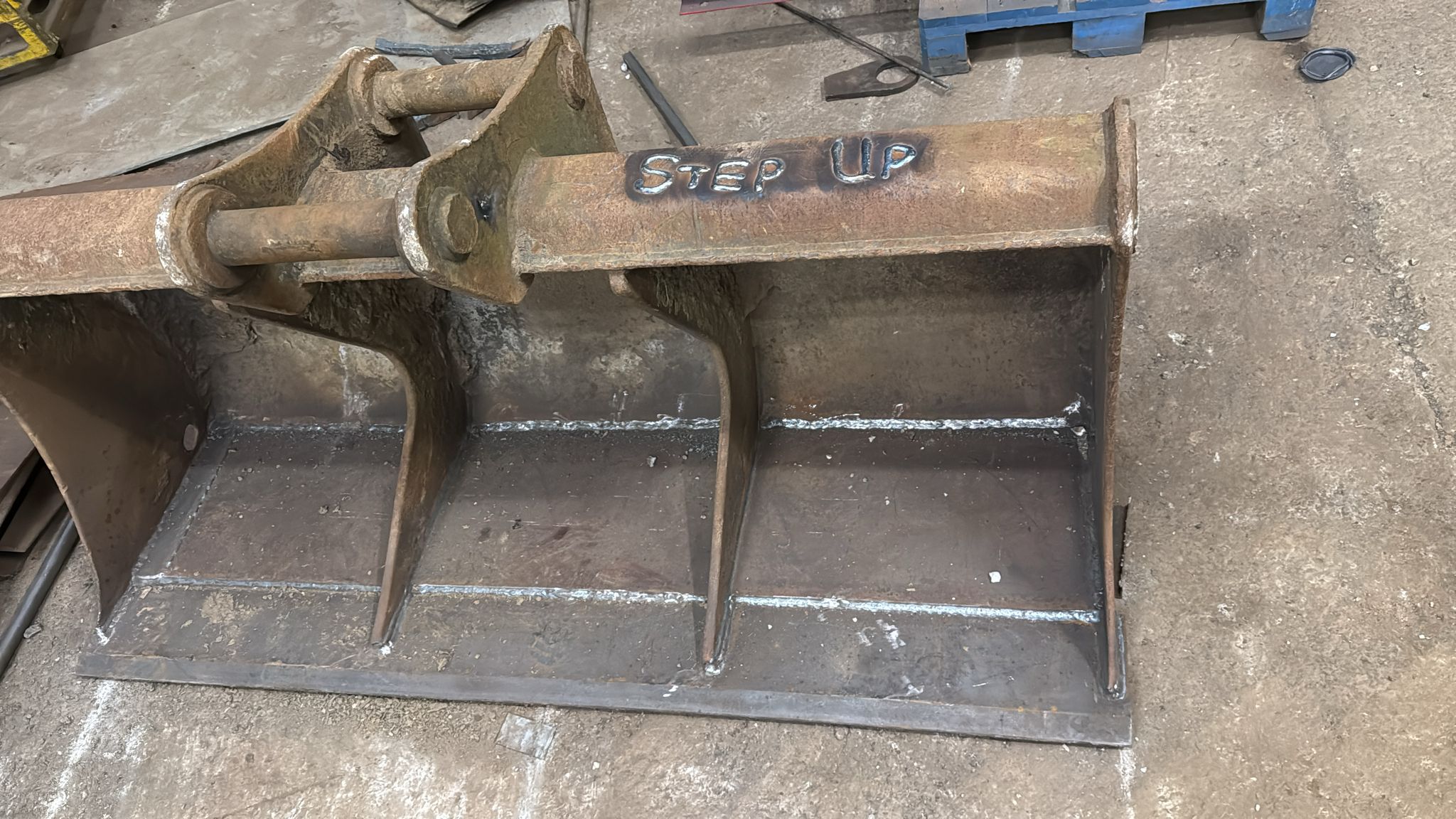 A rusted metal excavator bucket attachment lies on the ground, the words "STEP UP" expertly welded onto it using MOT Welding. The background features a concrete floor and part of a blue wooden pallet.