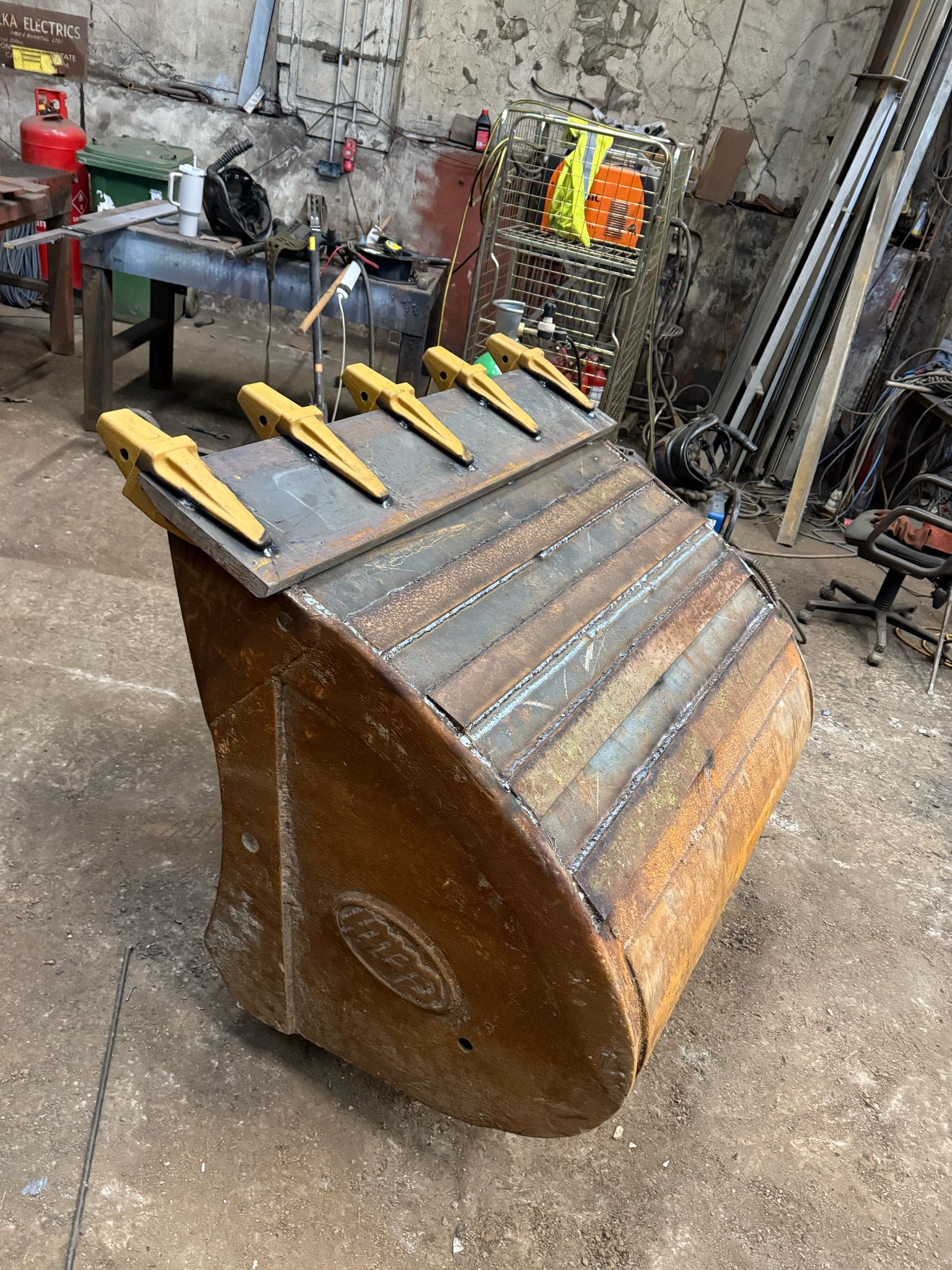 A large metal excavator bucket rests on a concrete floor in the workshop, freshly fitted with five new yellow teeth. In the background, tools, equipment, and metal bars are scattered against the industrial walls, ready for MOT welding or plant repair by our skilled mobile welder.