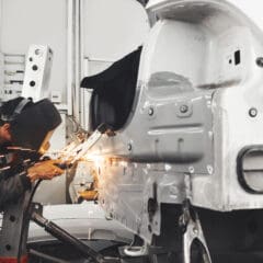 A worker in protective gear expertly welds a metal car body panel in a garage, creating bright sparks. The scene, reminiscent of an MOT welding operation, is set against an industrial backdrop filled with machinery and tools.