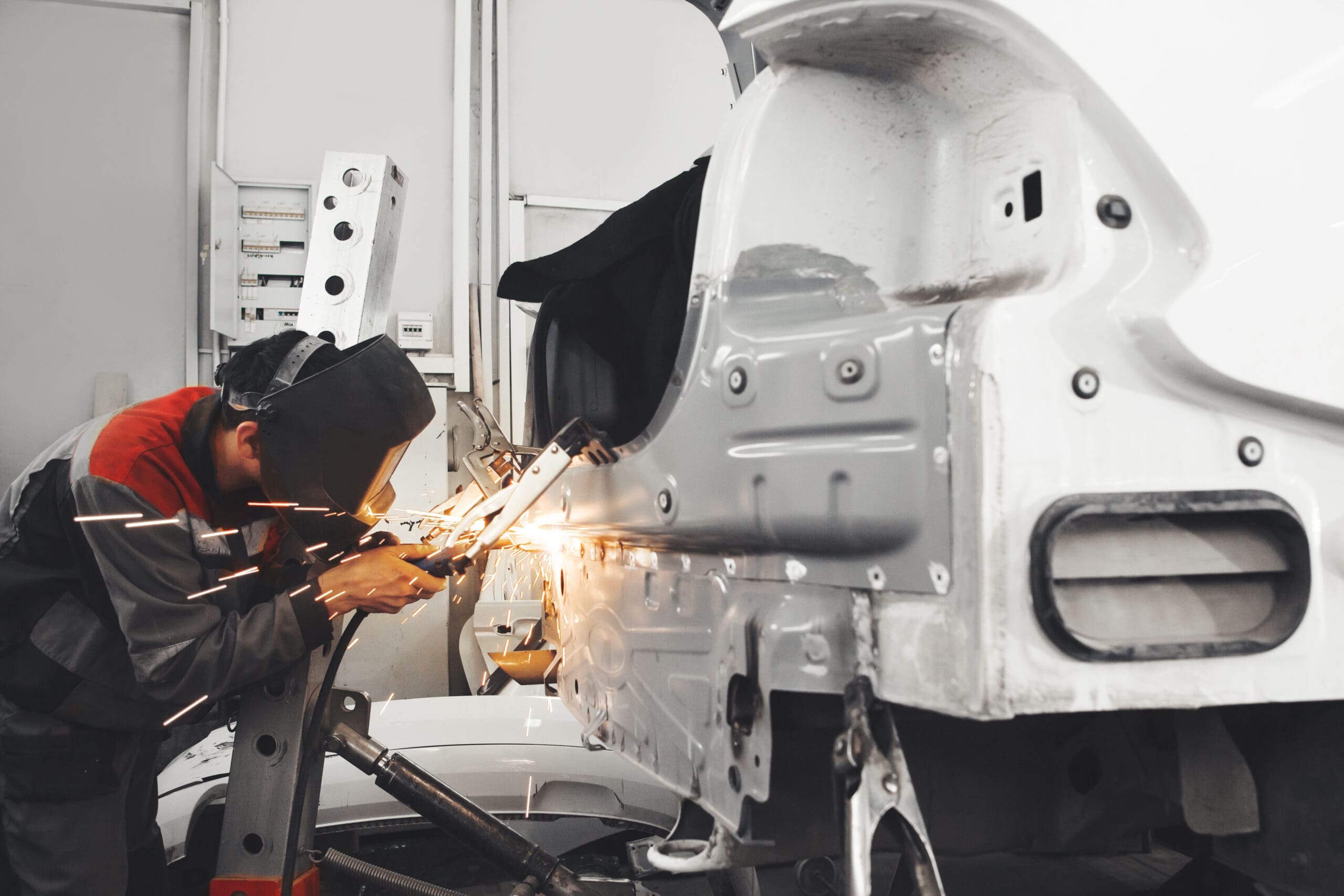 A worker in protective gear expertly welds a metal car body panel in a garage, creating bright sparks. The scene, reminiscent of an MOT welding operation, is set against an industrial backdrop filled with machinery and tools.