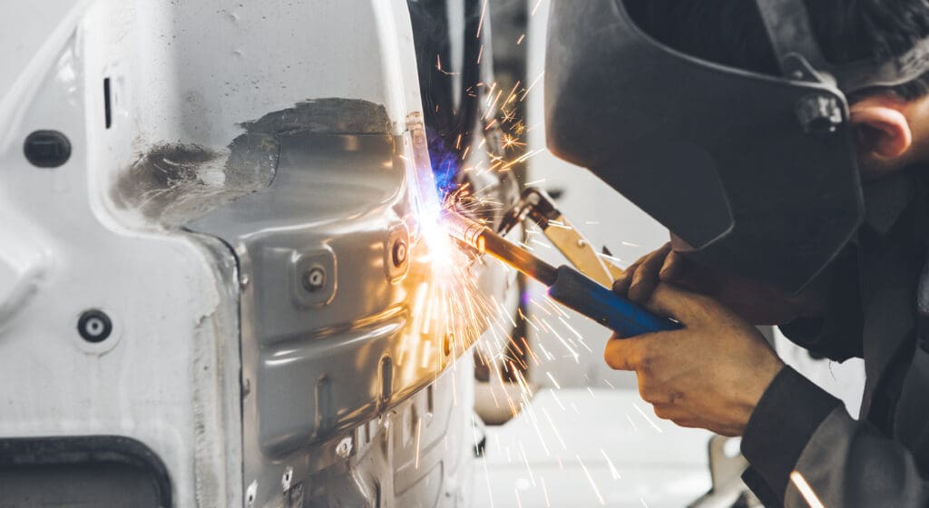 A mobile welder, wearing a welding helmet, is expertly welding a metal car part. Sparks illuminate the vehicle's body as they focus on an area with visible welding marks. The industrial workshop setting highlights their specialized MOT welding skills.