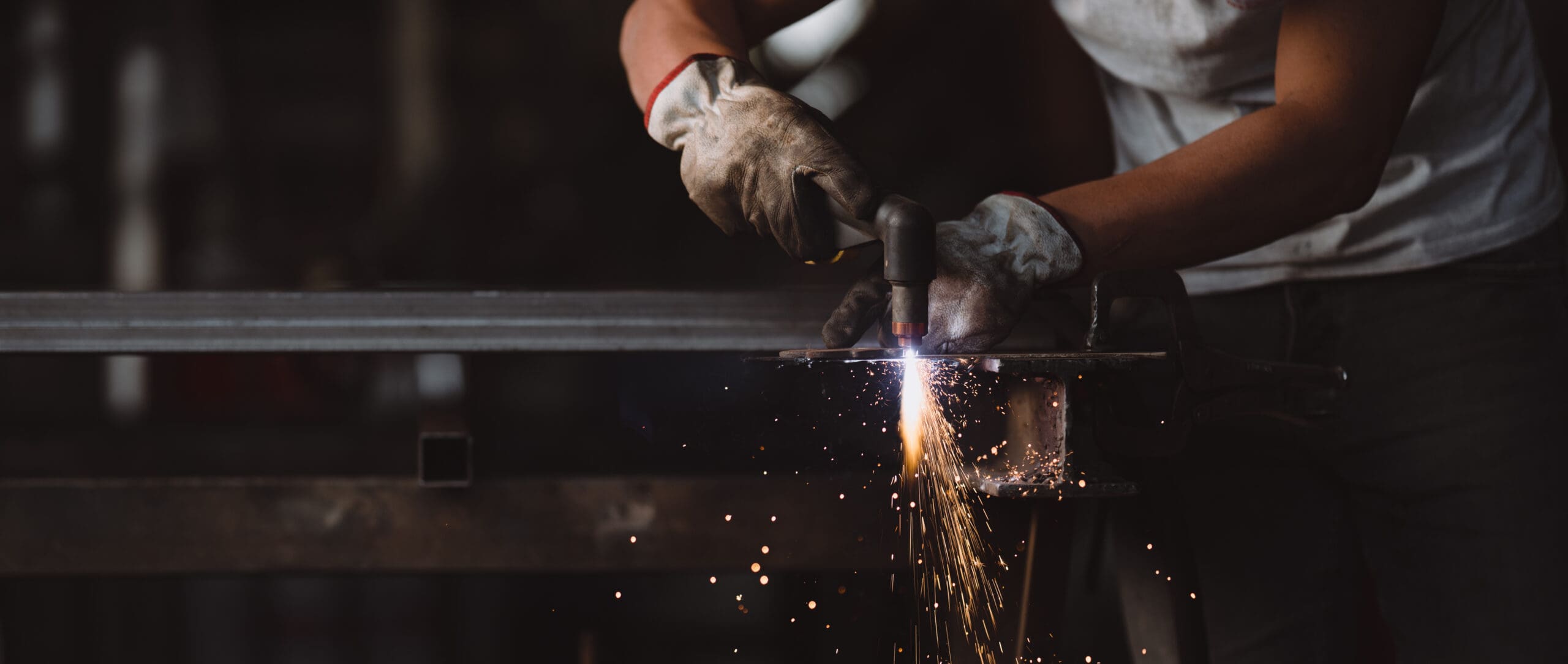 A mobile welder, clad in gloves, expertly wields a plasma cutter on a metal piece, sending sparks flying. The background is blurred to emphasize the focus on the precision and skill involved in this plant repair task.