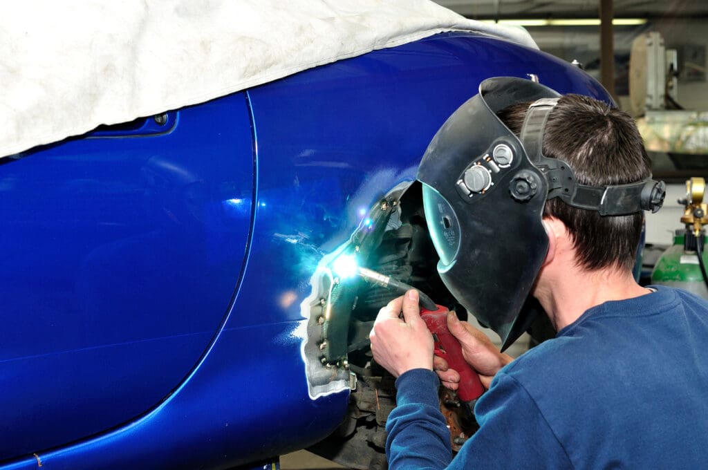 A mobile welder wearing a welding helmet works on the body of a blue car, creating bright sparks. The area is covered with a white tarp, and various tools and equipment are visible in the background.