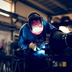 In an industrial workshop, a person wearing a protective helmet and gloves is performing MOT welding. Sparks fly as they expertly work on metal, surrounded by machinery and equipment. The well-lit space highlights the precision of the welding process.