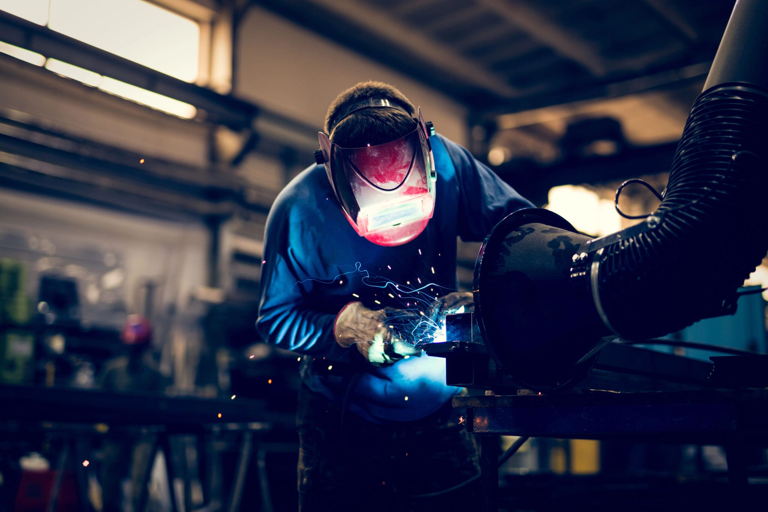 In an industrial workshop, a person wearing a protective helmet and gloves is performing MOT welding. Sparks fly as they expertly work on metal, surrounded by machinery and equipment. The well-lit space highlights the precision of the welding process.