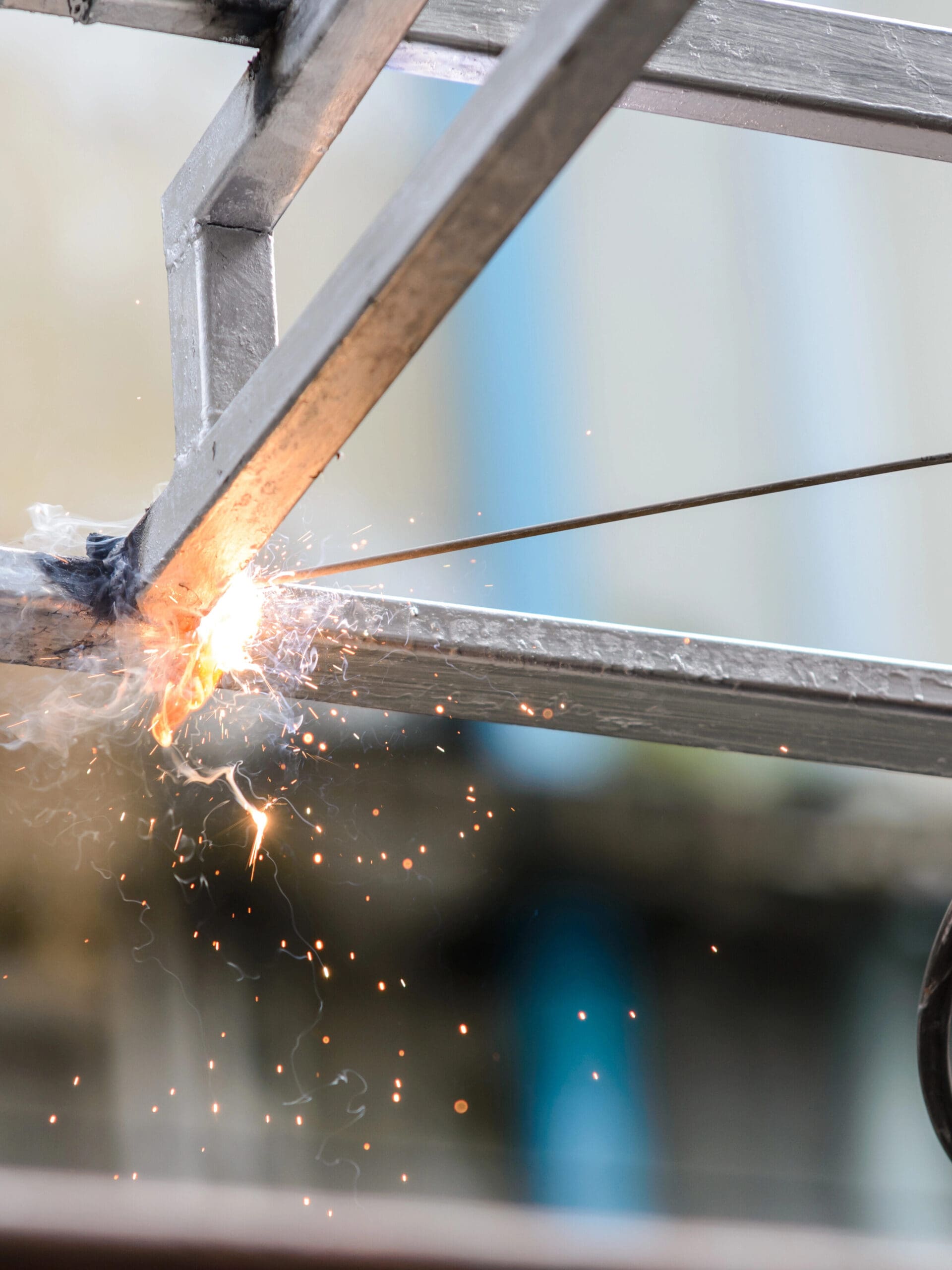 Close-up of a welding process showing bright sparks as a mobile welder expertly attaches a metal rod to a rectangular frame. The softly blurred background draws attention to the precise action, demonstrating skills often used in MOT welding and plant repair.
