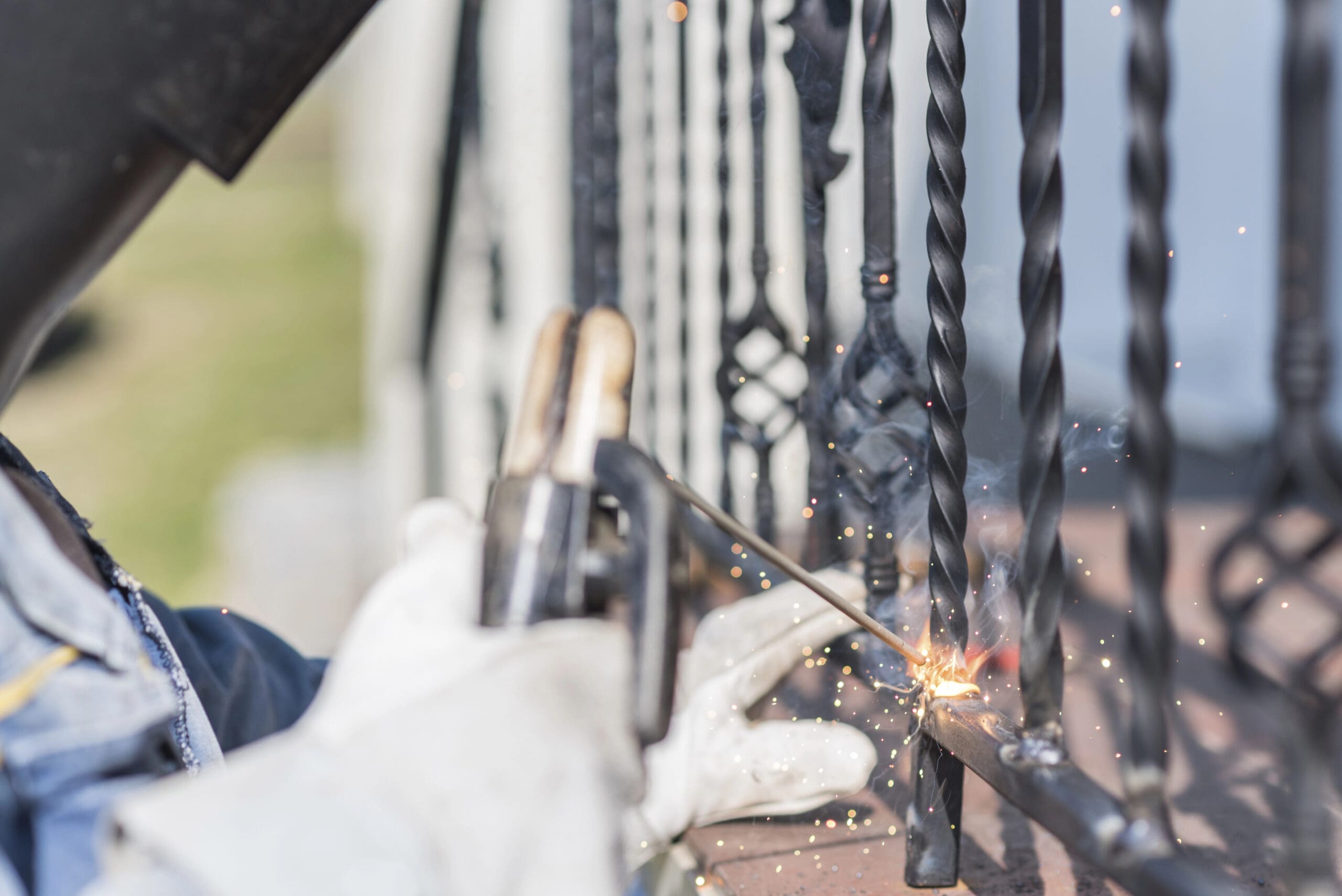A mobile welder in protective gear uses a welding torch on decorative metal railings, bright sparks flying from the point of contact. The scene unfolds outdoors, highlighting the precision and skill involved in the welding process.