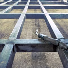 A metallic framework structure lies on a concrete floor, ready for a mobile welder's touch. A welding torch rests on one of the beams, hinting at ongoing MOT welding work. The framework's intersecting rectangular metal bars form a glowing grid pattern under bright lights, emphasizing its gleaming surface.