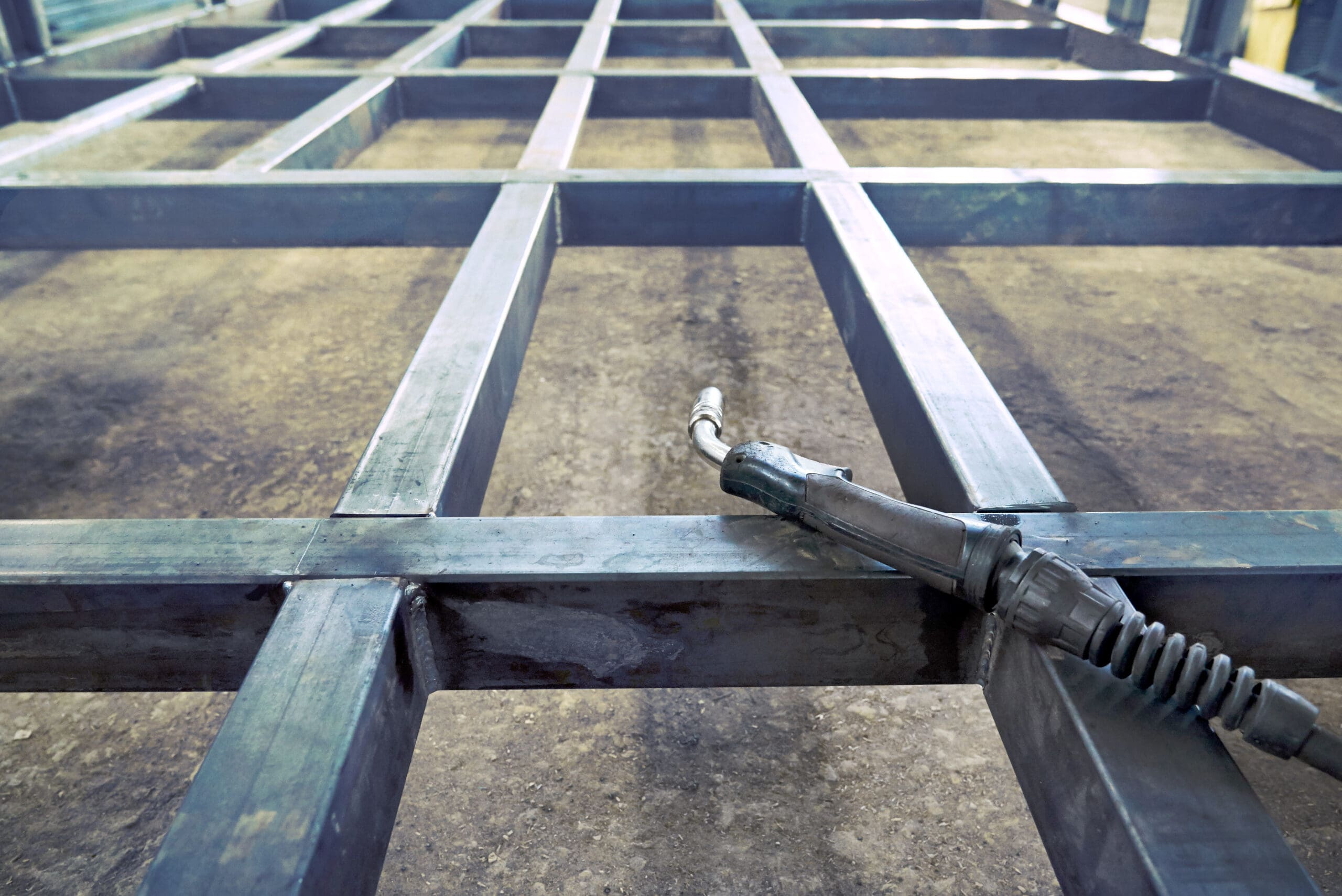 A metallic framework structure lies on a concrete floor, ready for a mobile welder's touch. A welding torch rests on one of the beams, hinting at ongoing MOT welding work. The framework's intersecting rectangular metal bars form a glowing grid pattern under bright lights, emphasizing its gleaming surface.