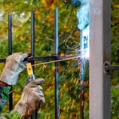 Two workers wearing protective gloves and helmets are expertly welding metal bars on a gate, showcasing their skills as mobile welders. Sparks fly from the process, and a bluish light is emitted, all set against a backdrop of blurred green foliage.