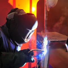 A welder in a protective helmet and gloves works in a dimly lit workshop, expertly executing MOT welding. Bright sparks fly from the tool as he joins metal pieces. The background is warm with orange and red hues, casting a glow around the dedicated mobile welder.