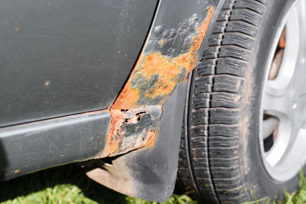 Close-up of a car with significant rust damage on the lower side panel next to the wheel. The metal is corroded, showing orange and brown discoloration, with some holes present. A mobile welder can address these issues before your next MOT check. The tire and grassy ground are partially visible.