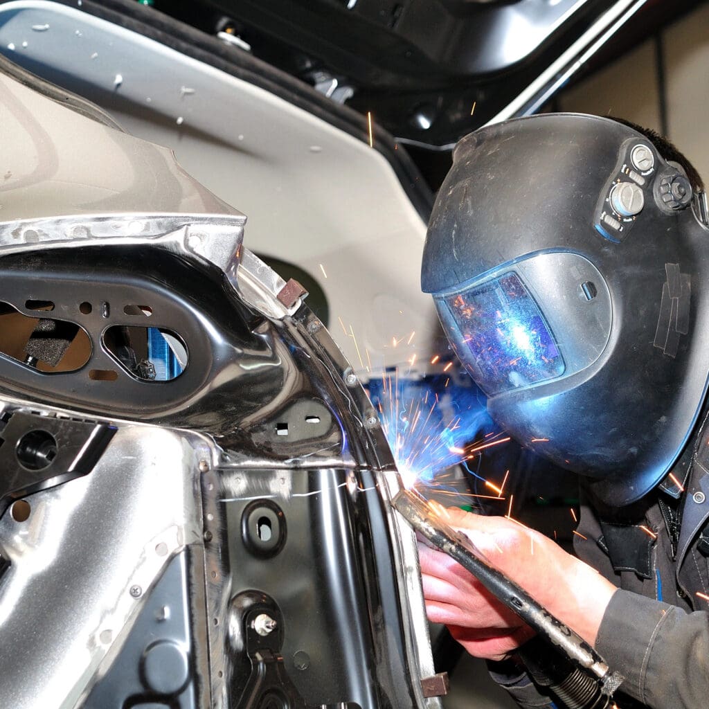 A mobile welder wearing a protective mask works diligently on a metal frame, creating bright sparks. Focused on joining two sections of metal, the welder is surrounded by tools and machinery, perfect for plant repair or MOT welding needs.