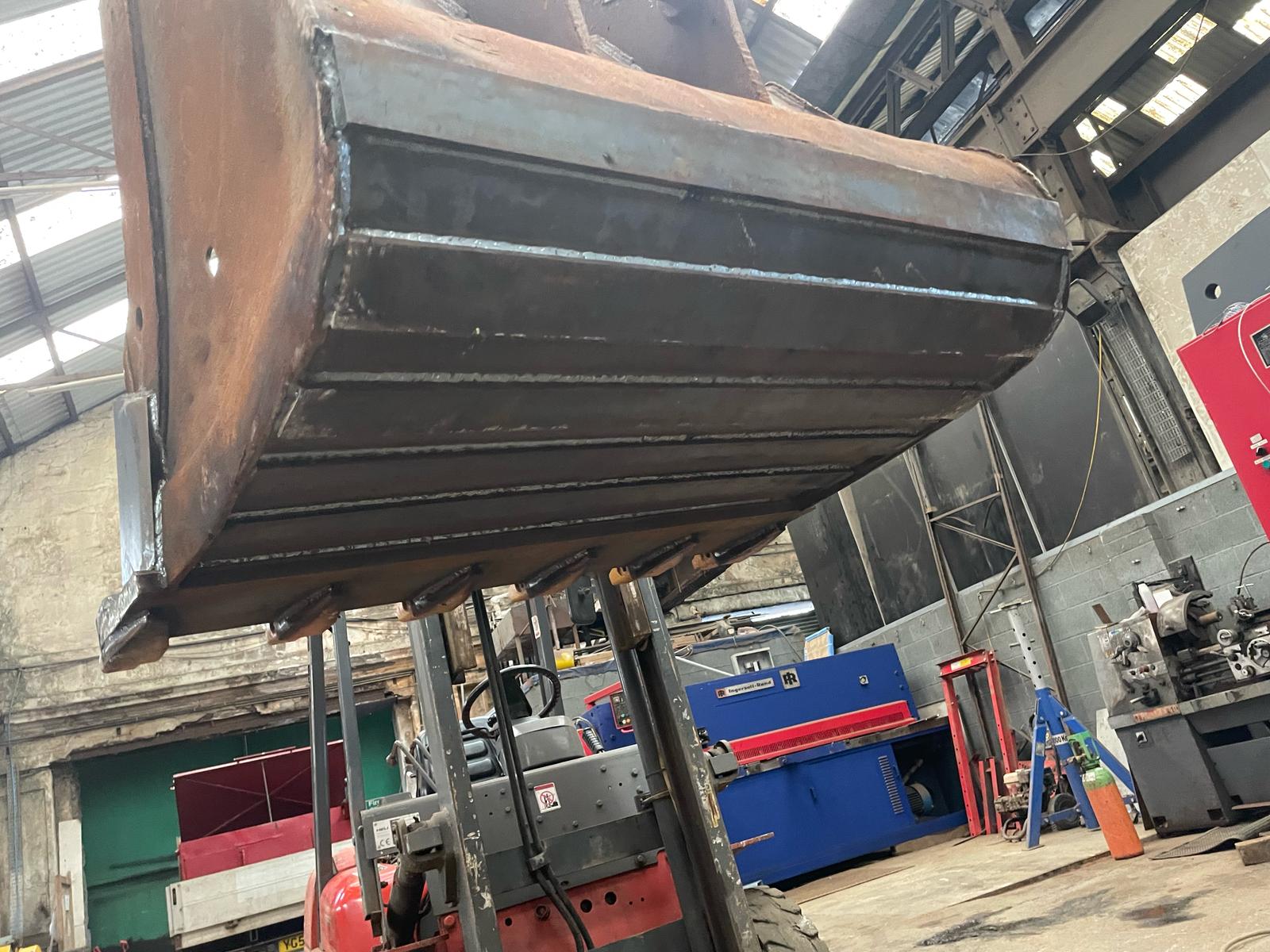 A large metal industrial machine part is being lifted by a forklift inside the workshop, surrounded by tools and structural beams. Natural light streams in through the partially visible roof, creating an ideal setting for MOT Welding amid diverse plant repair equipment.