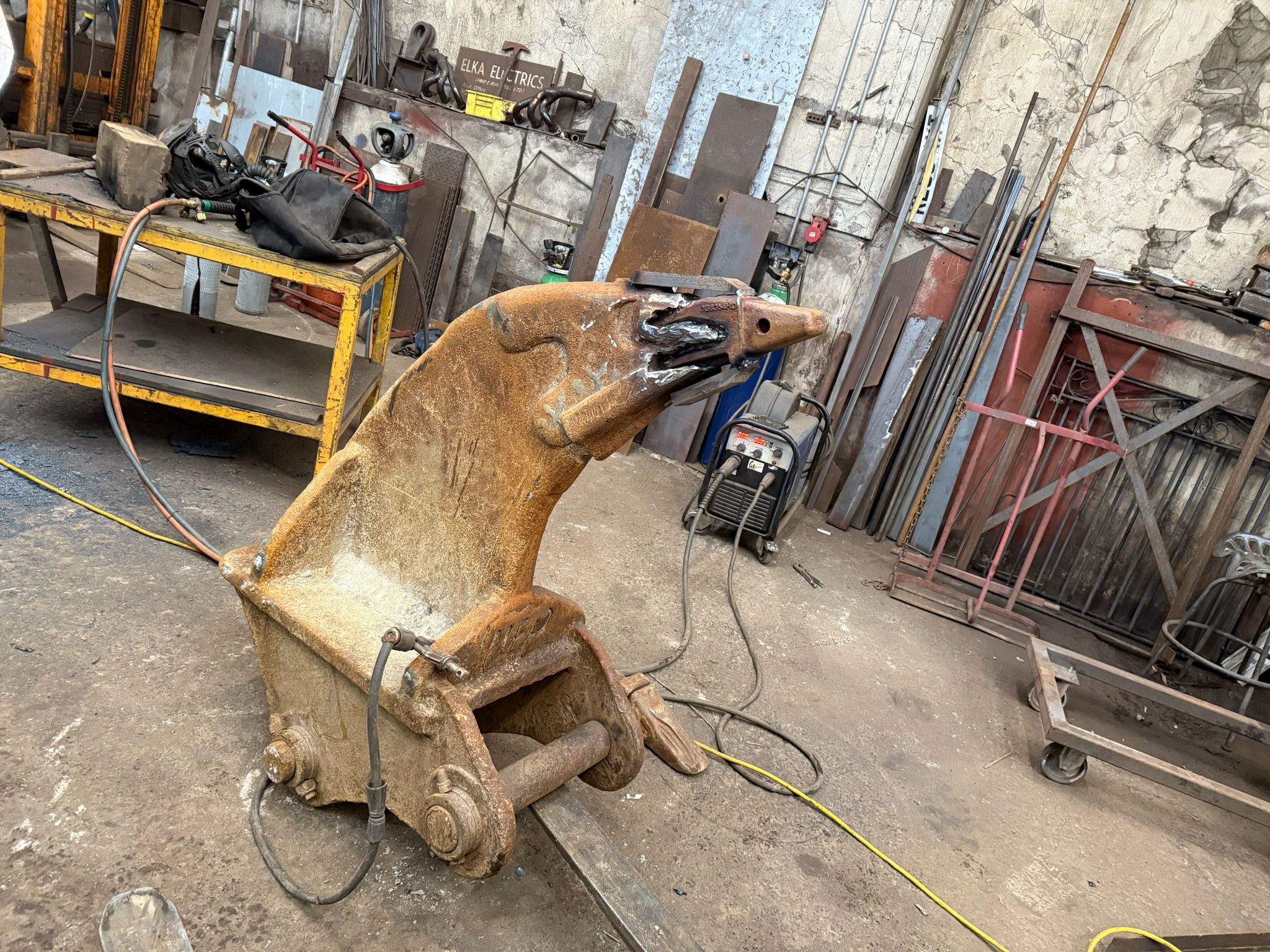 A large rusty metal bucket attachment sits on the floor of an industrial workshop, surrounded by tools and equipment for plant repair. The walls are worn and the space appears cluttered with various cables and mobile welder gear visible.