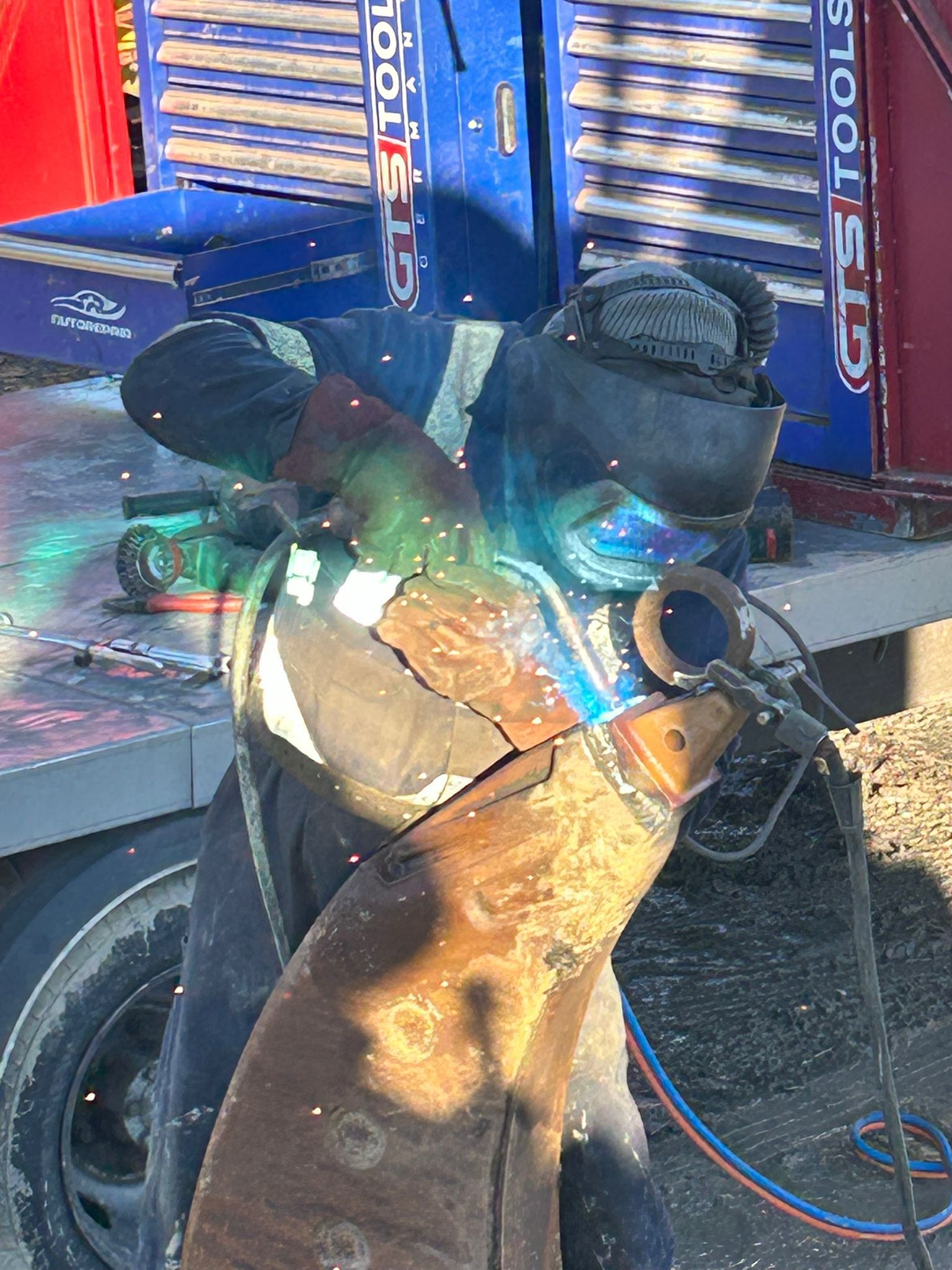A mobile welder in protective gear uses a grinder on a large, rusty metal piece in front of a toolbox. Sparks fly as the outdoor sun illuminates the plant repair scene.