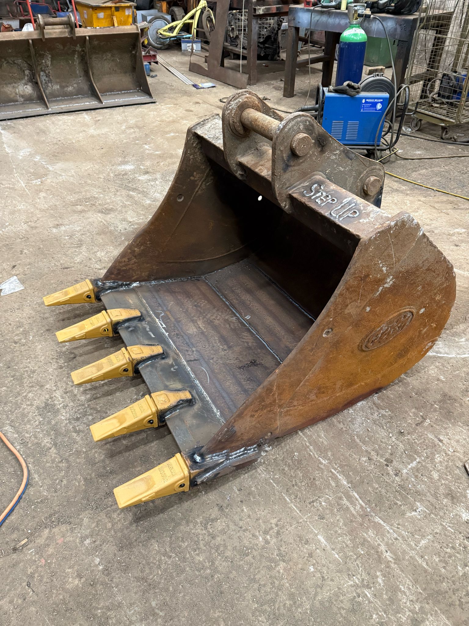 A heavy-duty excavator bucket with four yellow teeth sits on a concrete floor in an industrial workshop, surrounded by various tools and machinery. A mobile welder stands ready for MOT welding tasks or plant repair, a testament to the shop's versatile capabilities.