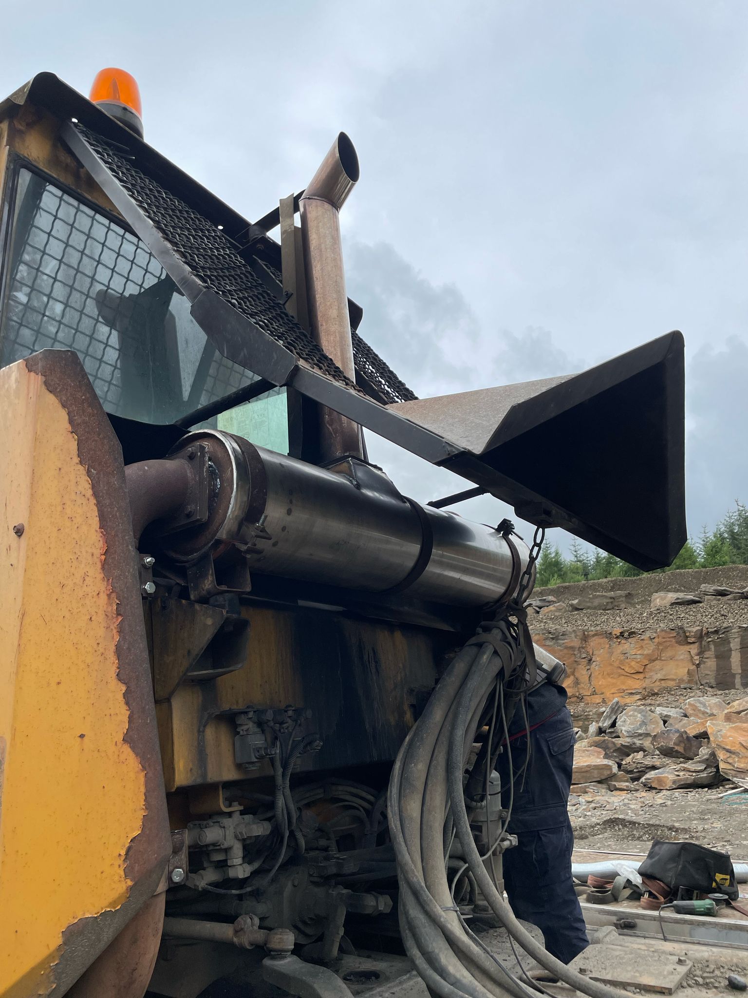 A large construction machine in a quarry shows signs of wear with its metal and hoses. An individual in dark clothing, possibly doing plant repair or MOT welding, is partially visible as they work on the equipment. The sky is overcast, casting a somber tone over the rocky terrain in the background.