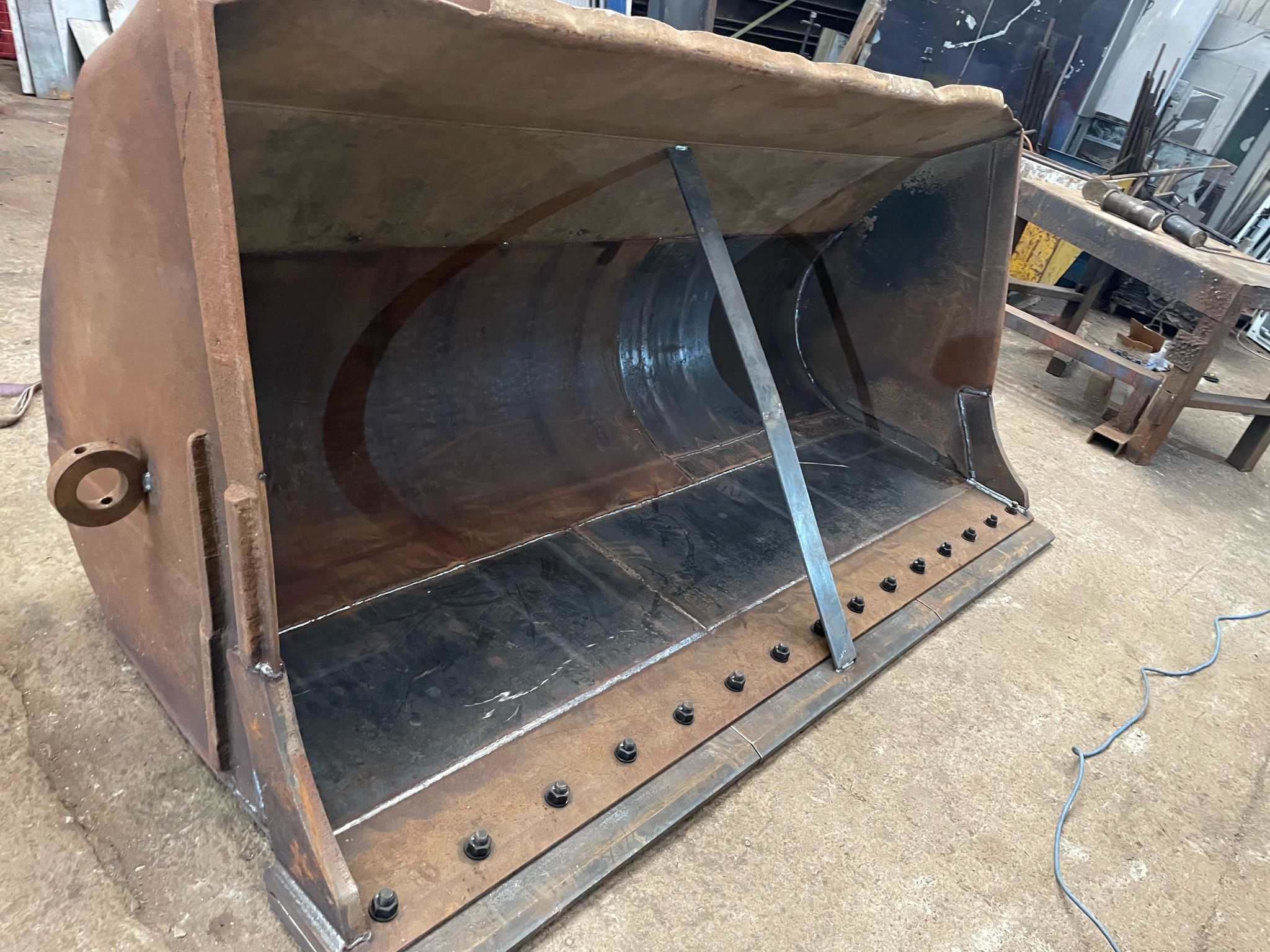 A large rusted metal bucket, possibly from construction equipment in need of plant repair, rests on a concrete floor. It's open, with bolts lining the bottom edge and a metal support bar inside. Various tools and equipment are visible in the background.