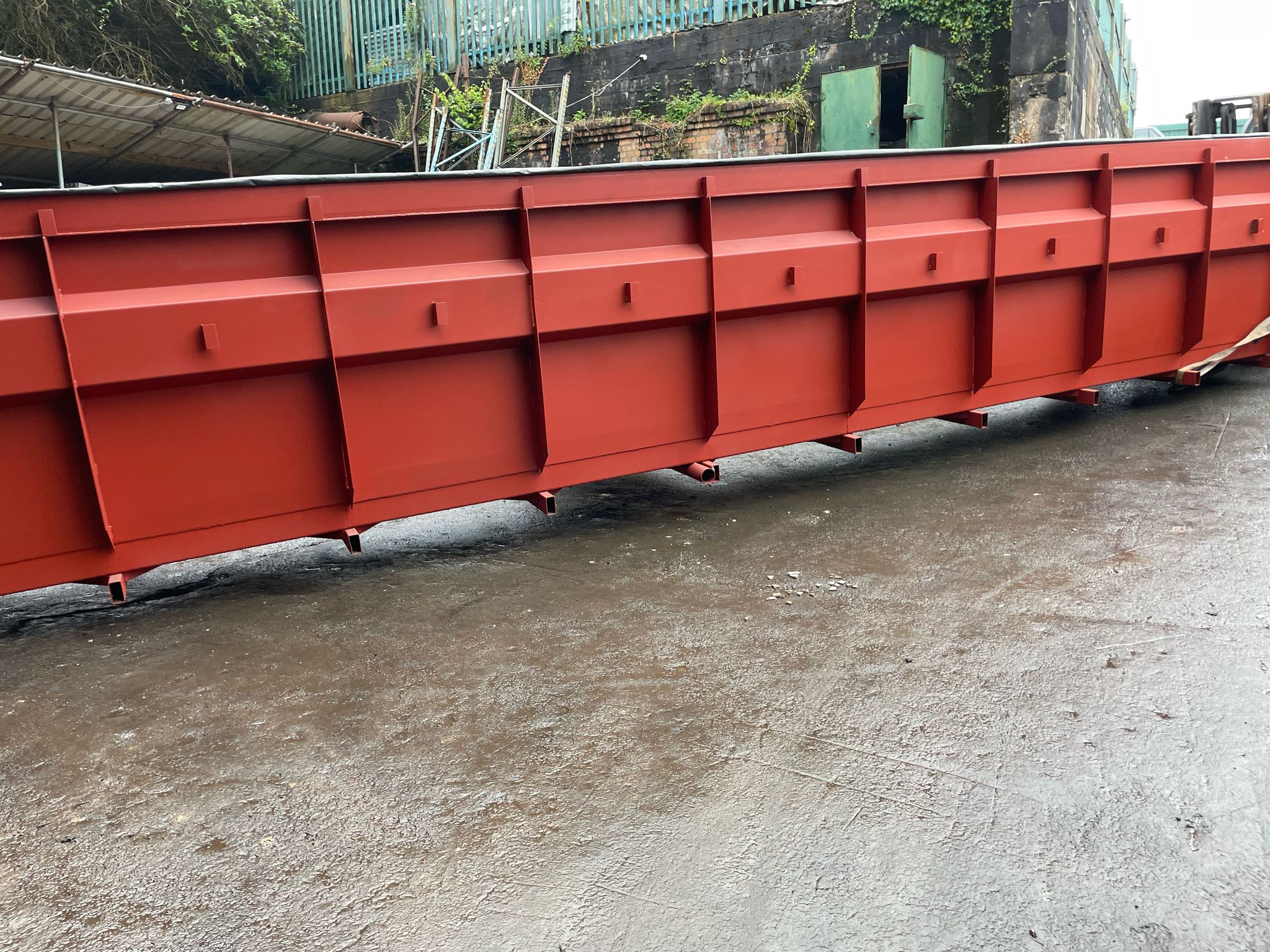 A large, red industrial container sits on a wet concrete surface. Adjacent to a site set for plant repair, the container features rectangular sections with reinforced edges. It's located in an outdoor area with metal fencing and greenery providing a natural contrast in the background.