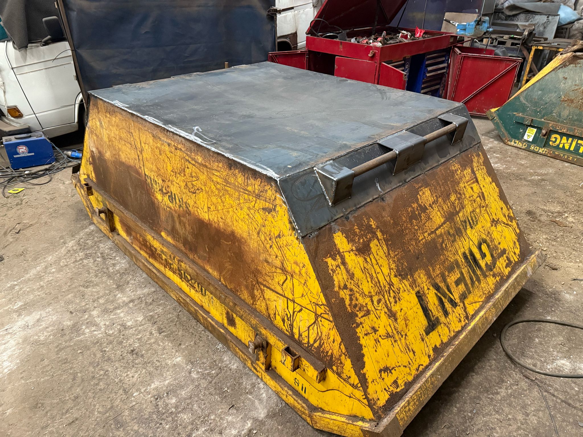 A rusty, yellow dumpster with a flat, modified metal lid sits in a workshop where mobile welders and tools suggest a busy industrial environment. Other vehicles hint at ongoing plant repairs and MOT welding tasks in the background.