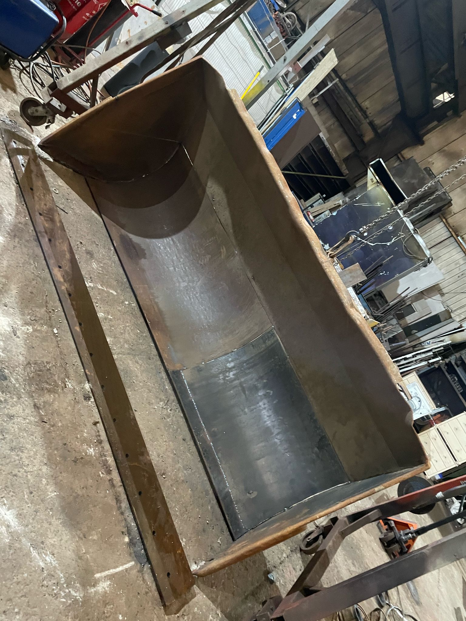 A large, empty metal container is positioned on a concrete floor in a bustling warehouse. Amidst the tools and machinery, there's a ladder and blue storage cabinet, hinting at ongoing plant repair. The setting exudes an industrial vibe typical of MOT welding operations.