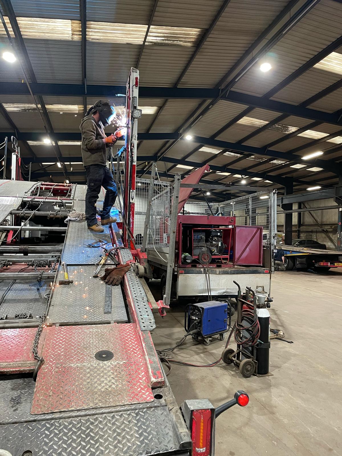 A mobile welder is diligently working on a large truck inside a spacious workshop. The area is equipped with various tools and machinery, all vital for plant repair, and the space is brightly lit by overhead lights.