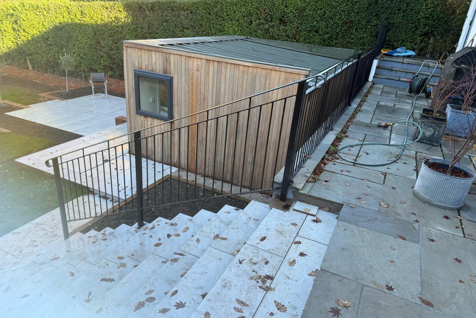 A backyard with a wooden shed, stone patio, and stairs leading down is surrounded by a metal railing. A mobile welder could be handy for repairs here. Nearby are a coiled hose and potted plants, with fallen leaves scattered on the ground.