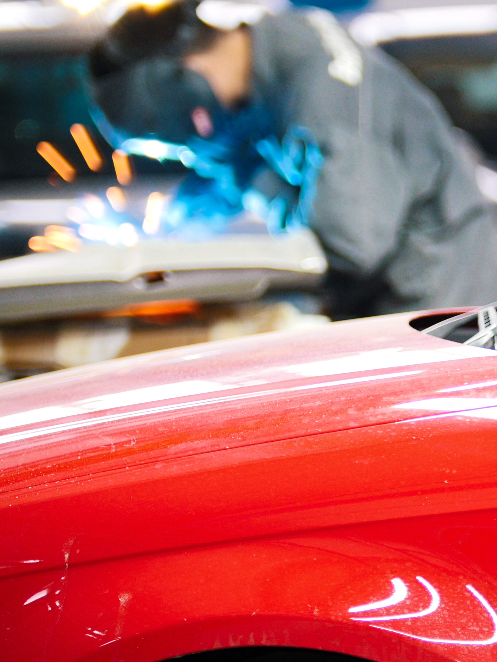 A mechanic in safety gear works on a car, generating blue sparks, in an automotive repair shop. A red car is prominently visible in the foreground.