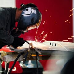 A person wearing a protective helmet and gloves is welding metal on a workbench. Bright sparks fly as the welding tool makes contact with the metal. The background is out of focus, showing a vivid red and dark blue environment.