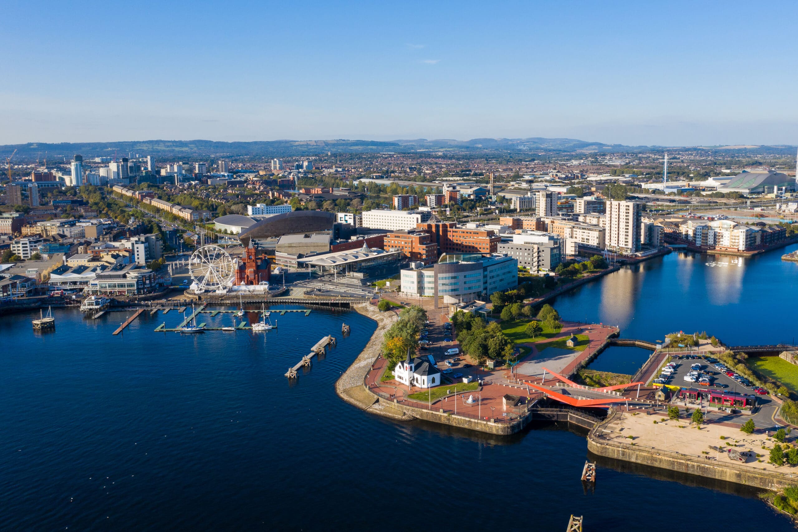 Aerial view of a waterfront cityscape featuring a large bay, marinas with boats, and modern buildings. A Ferris wheel and bridges are visible. The land is lush with greenery, mountains in the background under a clear sky—an idyllic setting for mobile welders specializing in plant repair.