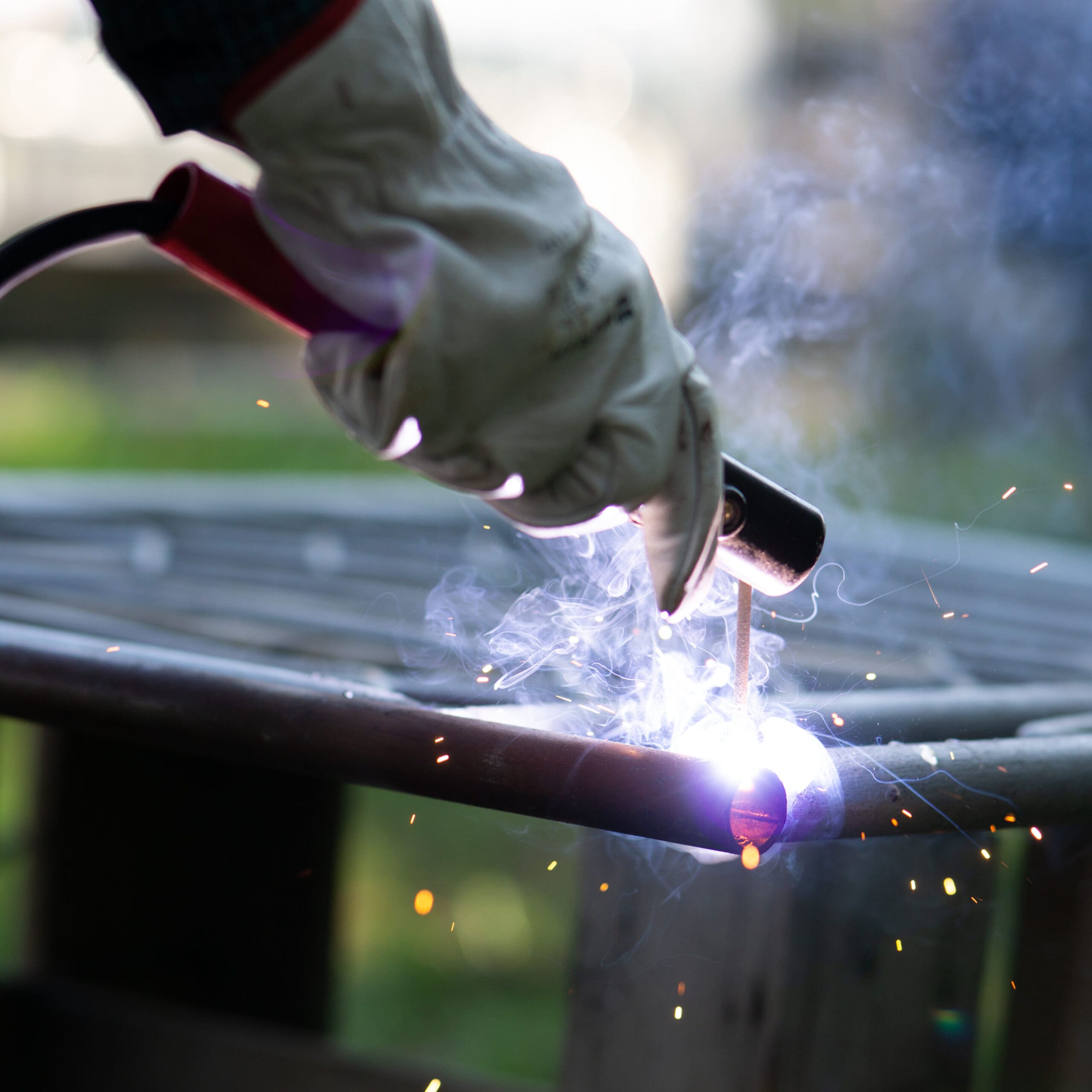 A person wearing protective gloves skillfully uses a welding torch on a metal pipe, creating bright sparks and smoke. As a mobile welder specializing in MOT welding, they work efficiently against a backdrop blurred with green and brown hues.